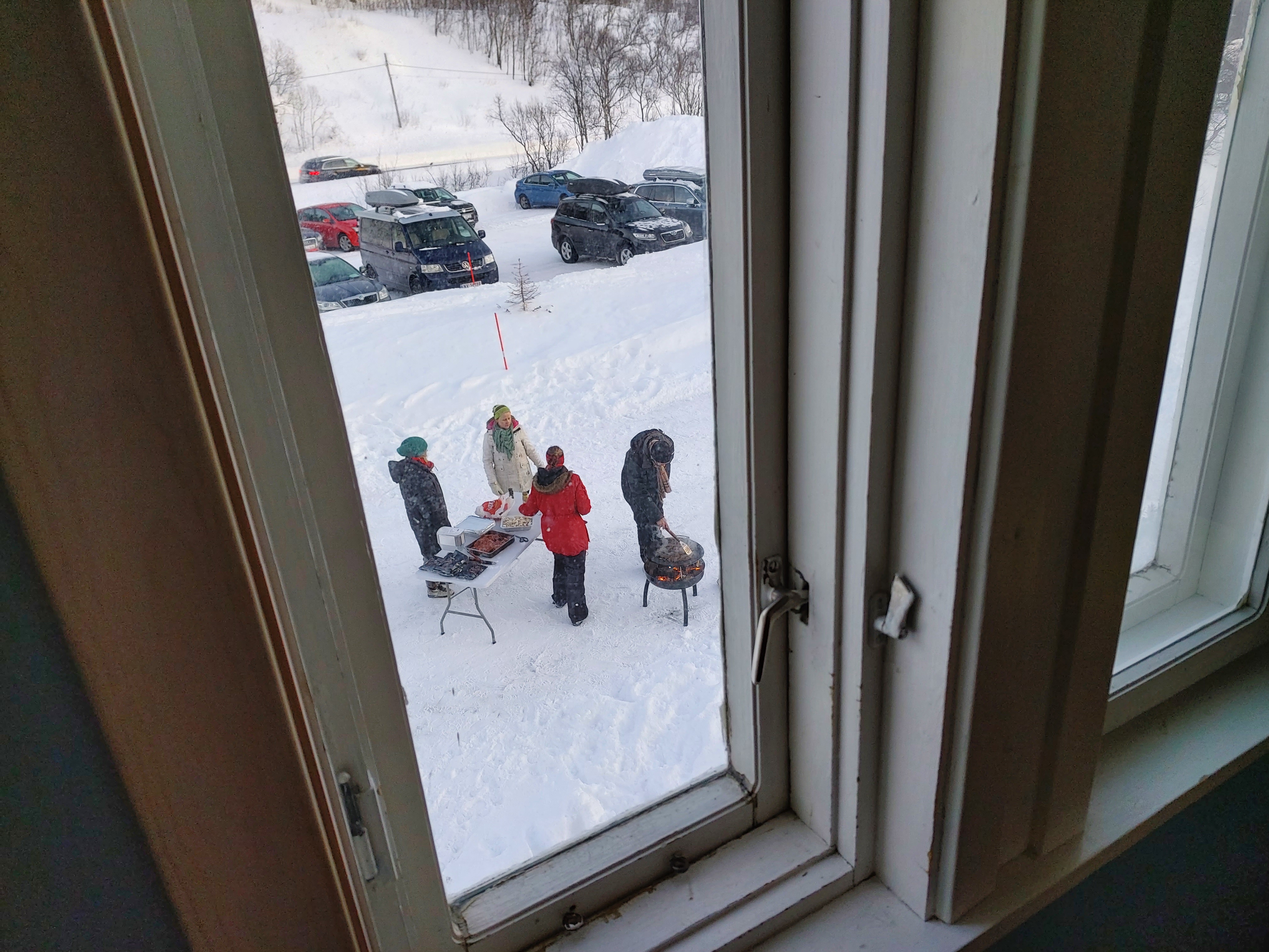 Bildet er tatt ut gjennom et vindu på galleriet i kirka, og vinduskarmene rammer inn motivet. Det står fire personer nede på bakken som er snøkledt, og litt lenger bort er det parkert flere biler. En person står lent over bålpanna og tre personer står ved et bord med mat på.